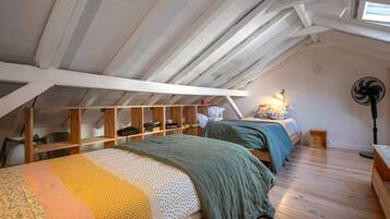 Une chambre à coucher aménagée dans un grenier, avec un plafond en pente, des poutres en bois et un puits de lumière. On y trouve deux lits avec une literie verte et jaune. Un ventilateur et une lampe sont posés sur le sol.