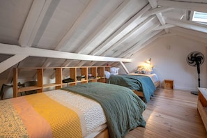A loft bedroom with a sloped ceiling, wooden beams, and a skylight. There are two beds with green and yellow bedding. A fan and a lamp are on the floor.