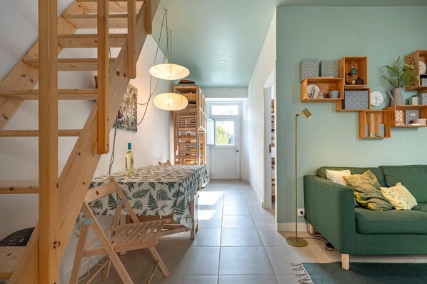 A room with a wooden staircase, a dining area with a table and chairs, a green sofa, and shelves with decorative items.