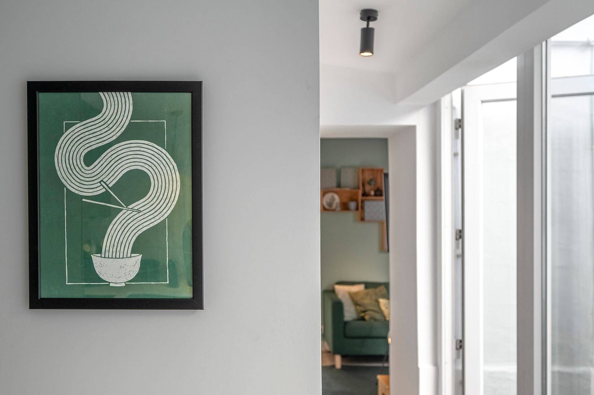Een lijstwerk aan de muur, een deuropening naar een kamer met een groene bank, en een plafondlamp.