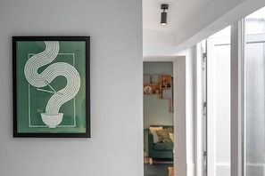 A framed artwork on a wall, a doorway leading to a room with a green sofa, and a ceiling light fixture.