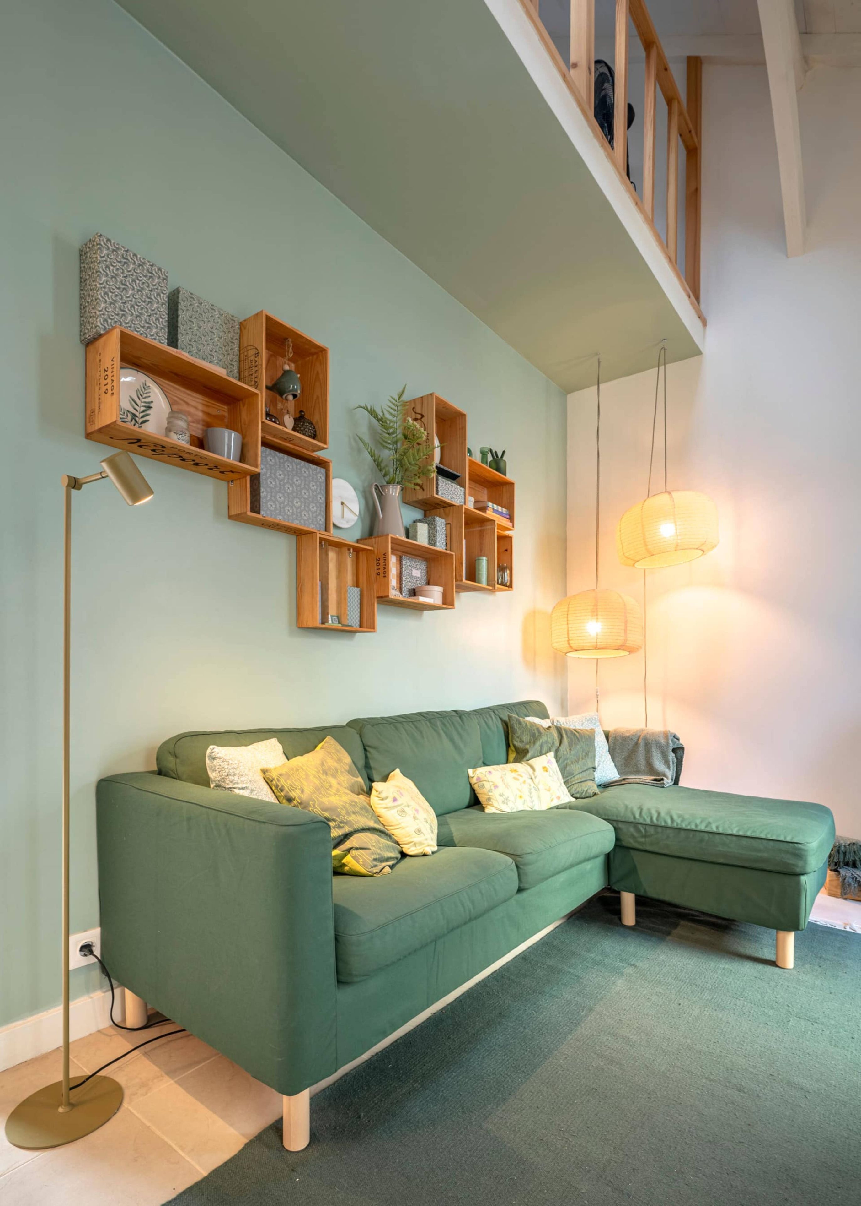 Un canapé vert agrémenté de plusieurs coussins, des étagères en bois au-dessus, sur lesquelles sont disposés divers objets, et une lampe sur pied.