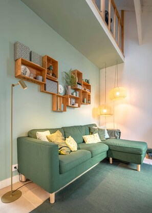 A green sofa with multiple cushions, wooden shelves above with various items, and a floor lamp.