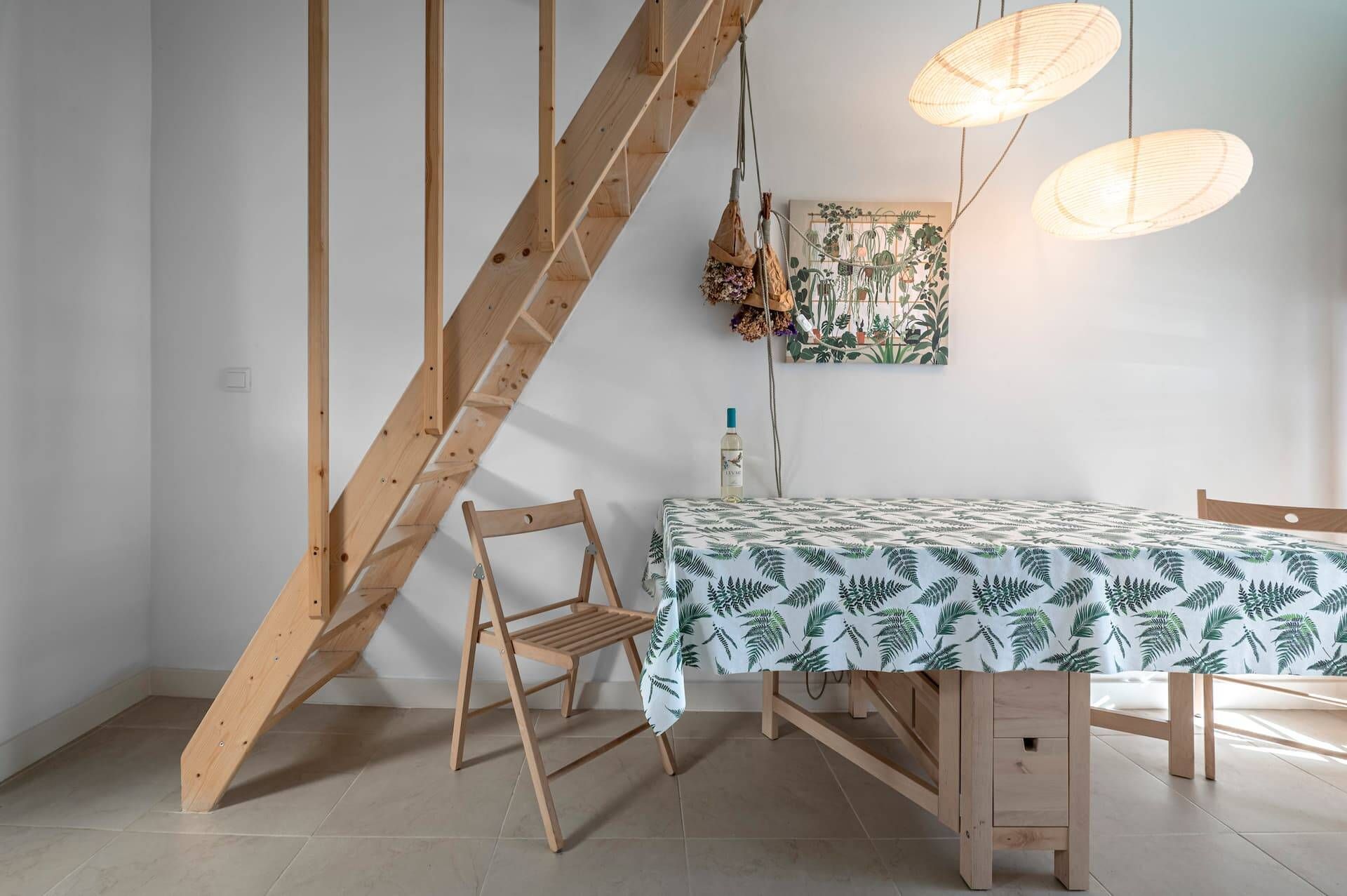 Un coin repas avec une table recouverte d’une nappe à motifs de feuilles, des chaises en bois, une échelle en bois et des luminaires suspendus.