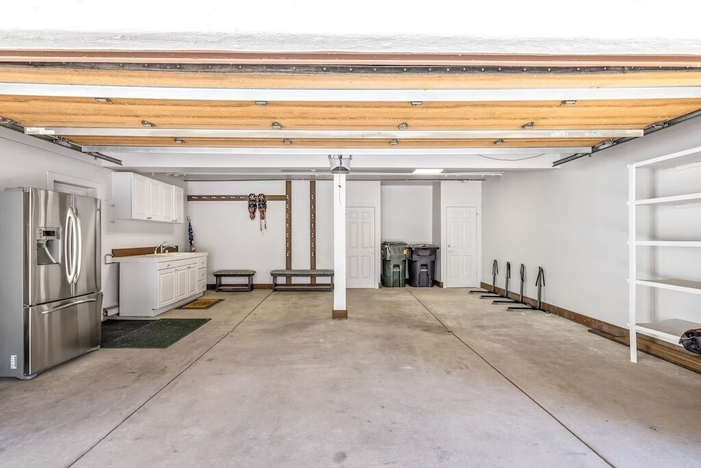 A spacious garage with a concrete floor, a stainless steel refrigerator, white cabinets, and a wooden ceiling.