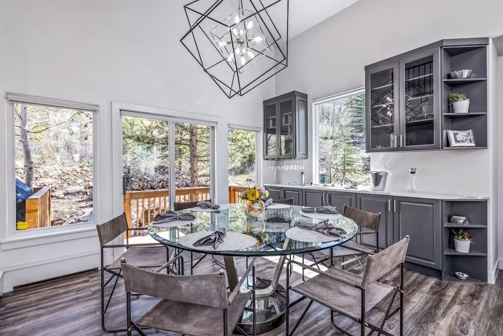 A modern kitchen with a glass dining table, dark cabinets, and large windows offering a view of the outdoors.