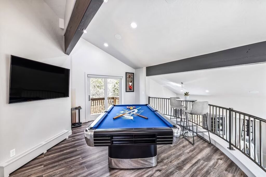 A pool table in a room with a balcony, a TV mounted on the wall, and a window with a view.
