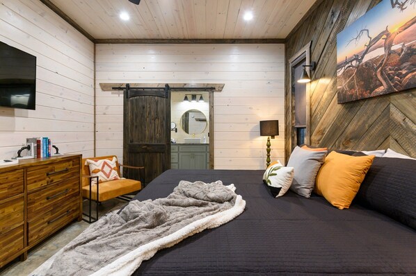A bedroom with a bed, a wooden dresser, a chair, a mirror, and a painting on the wall.