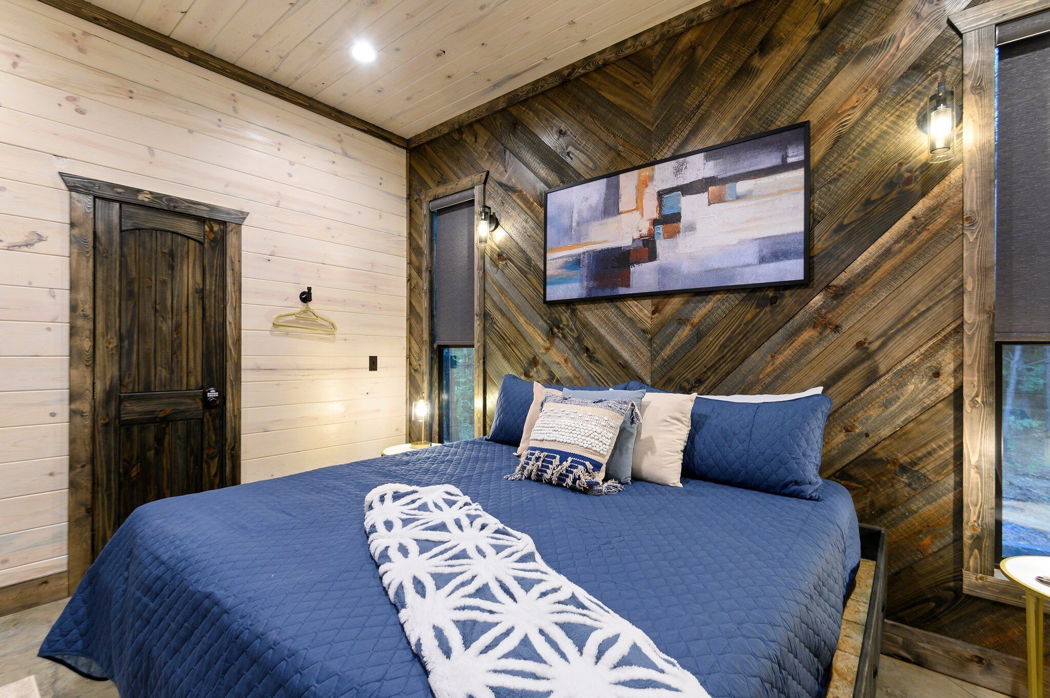 Un dormitorio con pared de madera en patrón de espiga, cama con ropa de cama azul, perchero y un cuadro sobre la cama.