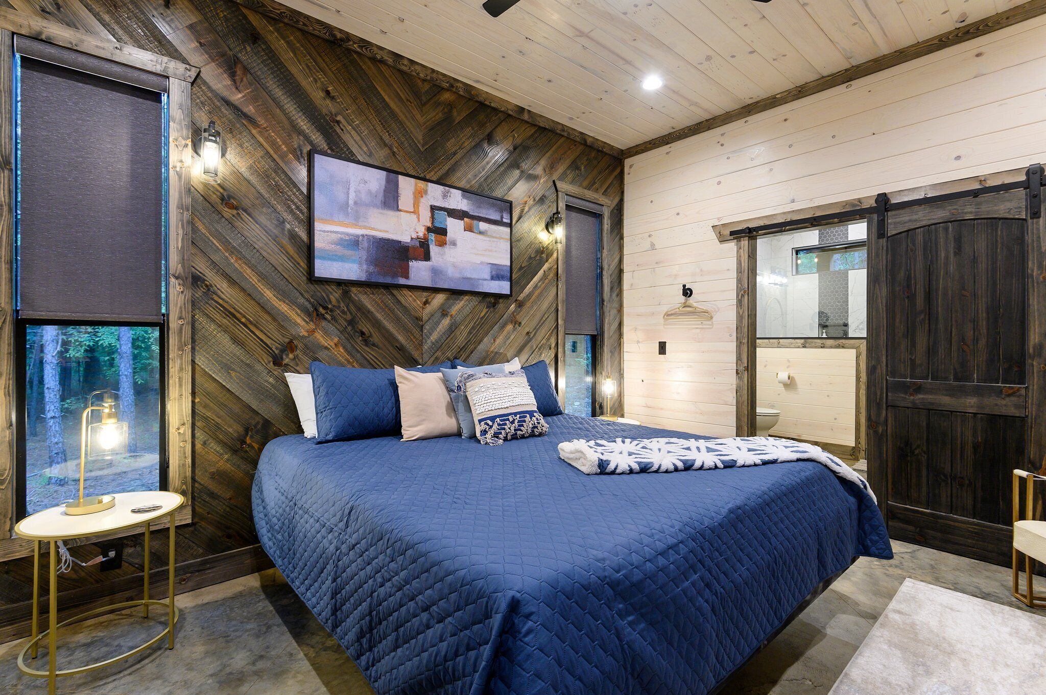 A bedroom with a large bed, a wooden wall, a painting, and a window with a view of trees.