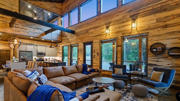 A spacious living area with a large sectional sofa, a coffee table, and a fireplace. There are multiple windows providing natural light and views of the outdoors. - Sip and Stay Hideaway (Broken Bow)