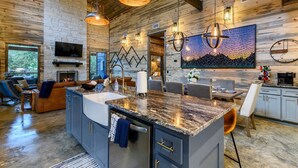 A spacious kitchen with granite countertops, a built-in oven, and a large island. There is a dining area with a table and chairs, and a seating area with a sofa and chairs. - Sip and Stay Hideaway (Broken Bow)