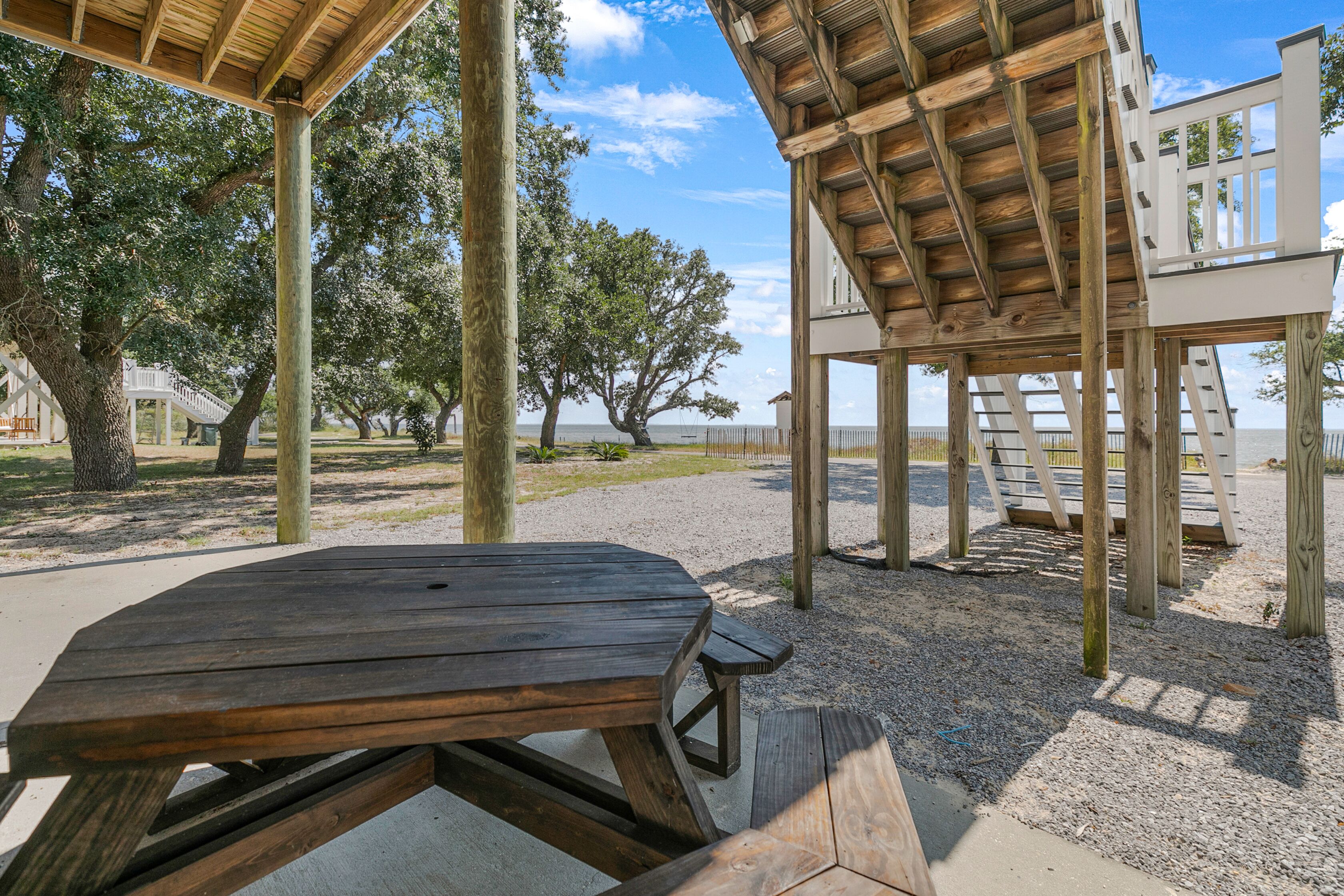 A wooden picnic table and a wooden deck structure with a white railing, surrounded by trees and a clear sky.