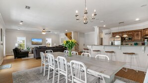 Een ruime woonkamer met een eettafel, witte stoelen, een kroonluchter en een keuken met houten kastjes en barstoelen.