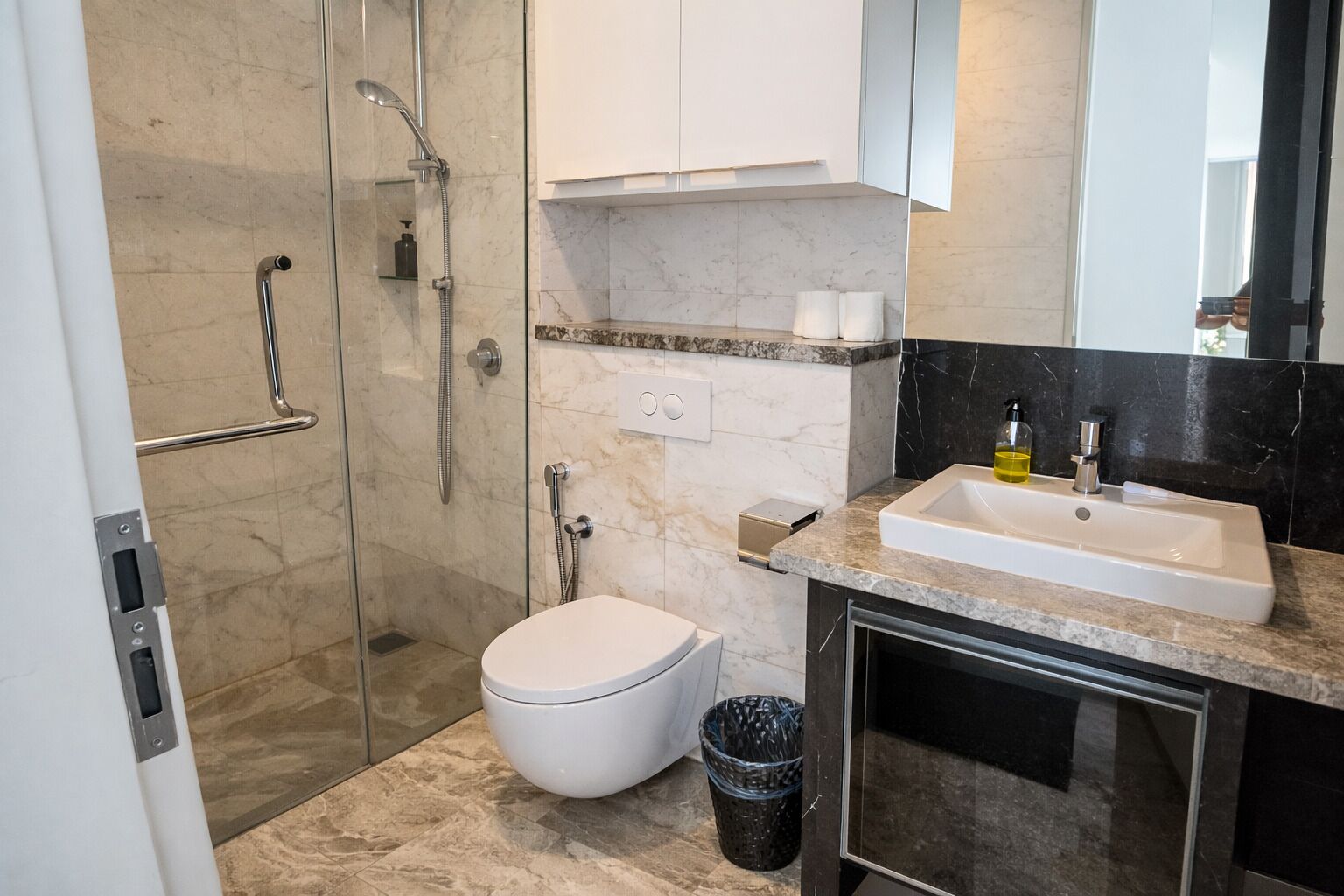 Un baño moderno con ducha de vidrio, inodoro, lavamanos con espejo y un cubo de basura.