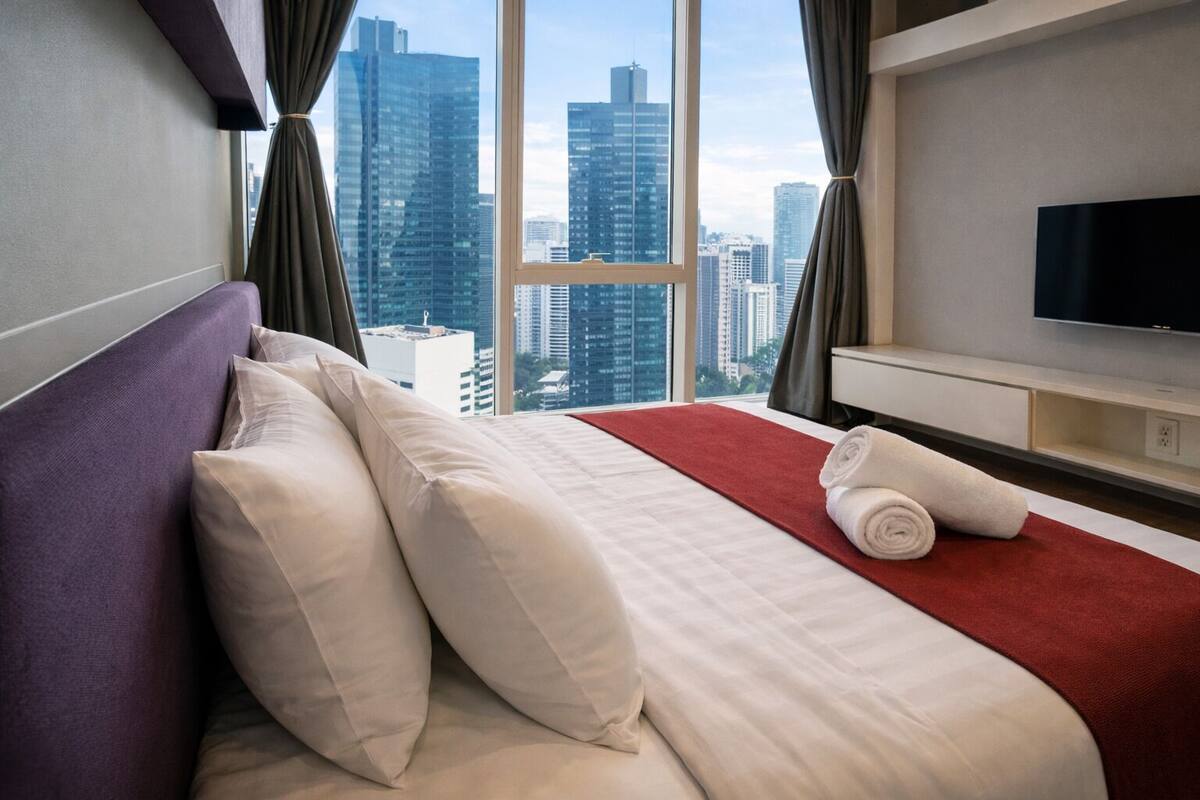 A hotel room with a large window, a bed with white linens, a red accent rug, and a flat-screen TV on the wall.