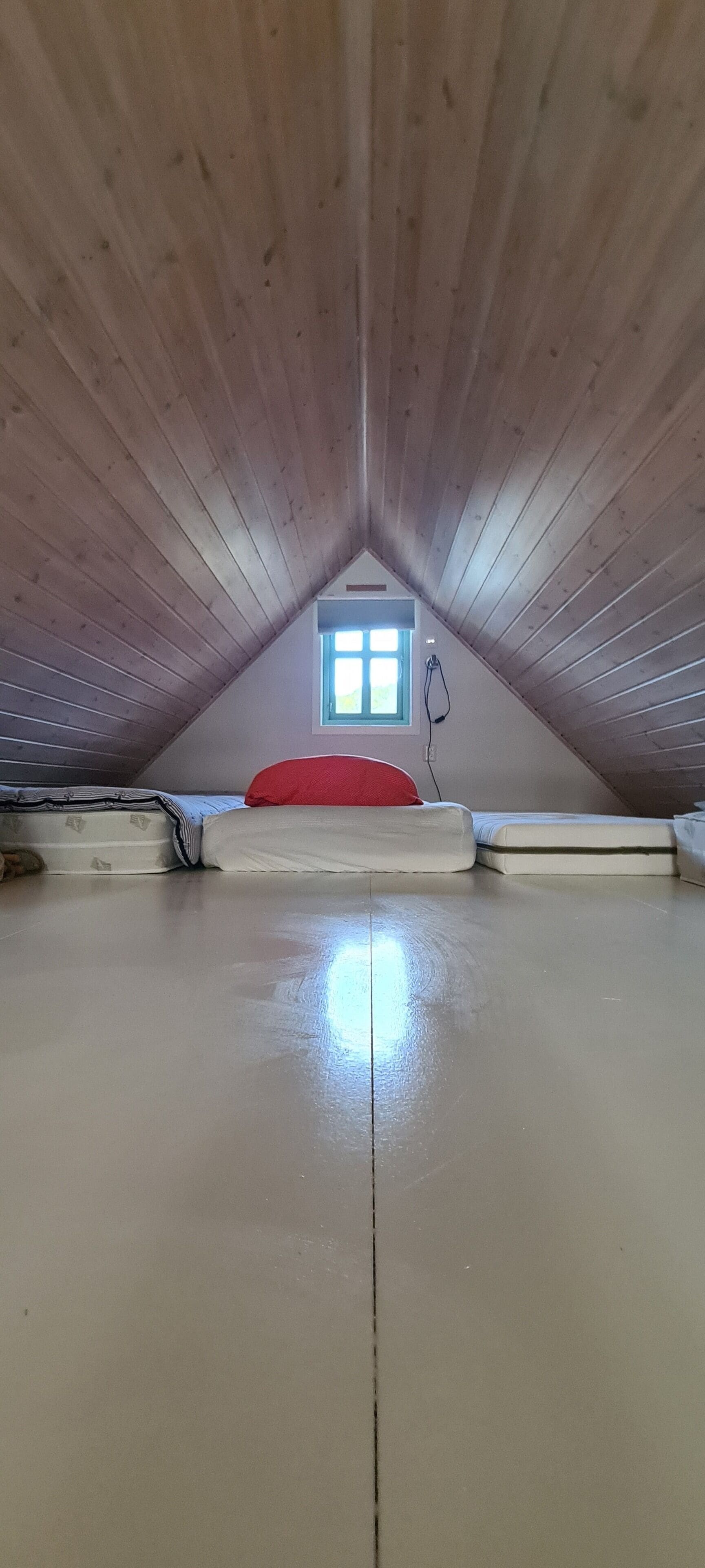 An attic room with a sloped wooden ceiling, a small window, and a red cushion on a mattress.