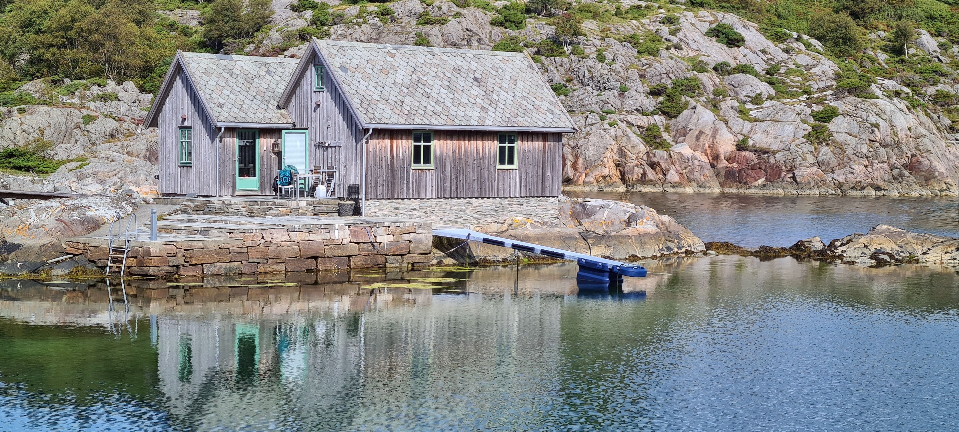 A wooden cabin with a stone dock extending into a calm lake, surrounded by rocky terrain.