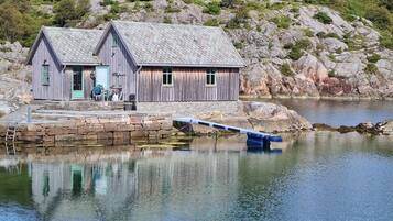A wooden cabin with a stone dock extending into a calm lake, surrounded by rocky terrain.