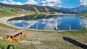 A lakeside view with a wooden picnic table, calm water, and a backdrop of mountains and houses.
