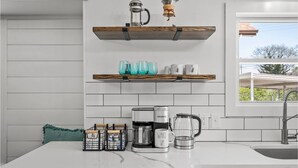 Une cuisine avec des meubles blancs, un plan de travail en marbre, une machine à café, une bouilloire et une étagère contenant de la verrerie.