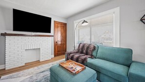 Un salon doté d’une cheminée en briques blanches, d’un canapé turquoise, d’une table basse en bois agrémentée d’un objet décoratif, d’un grand téléviseur à écran plat fixé au-dessus de la cheminée et d’une fenêtre avec des stores.