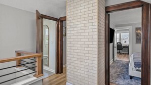 Un couloir avec une rambarde en bois, un mur en briques blanches et une porte donnant sur une chambre à coucher équipée d’un lit et d’un bureau.