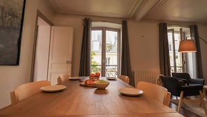 Een eetruimte met een houten tafel, stoelen, een fruitschaal en uitzicht op gebouwen door grote ramen.