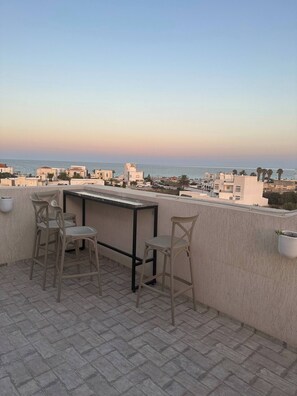 A terrace with two chairs and a table, overlooking a coastal cityscape with buildings and the sea. - Luxury Rooftop Wifi, Ac, Outdoor Barbecue (Mahdia)