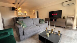 Een moderne woonkamer met een grijze bank, een groene fauteuil, een zwarte salontafel met kaarsen, en een flatscreen televisie waarop 'NETFLIX' staat.