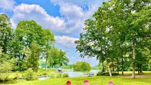 A circular seating area with red chairs around a central fire pit, surrounded by a grassy lawn with trees and a body of water in the background. - Lakefront Cabin Boat slip Wifi Fishing Firepit (Orange)