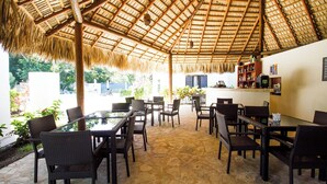 An outdoor seating area with wicker chairs and tables, a thatched roof, and a bar area with shelves of bottles.