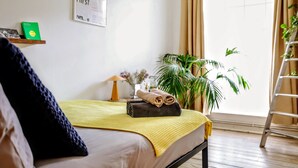 A neatly made bed with a yellow blanket, a rolled towel, and a small plant on the bedside table. There is a ladder leaning against the wall, a large window with curtains, and a framed picture on the wall. - Designer apartment in modern neighborhood - 15 minutes from central London (London)