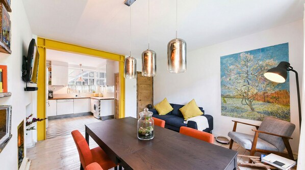 A modern living room with a dining table, sofa, and chairs. There is a kitchen in the background with a yellow accent wall.