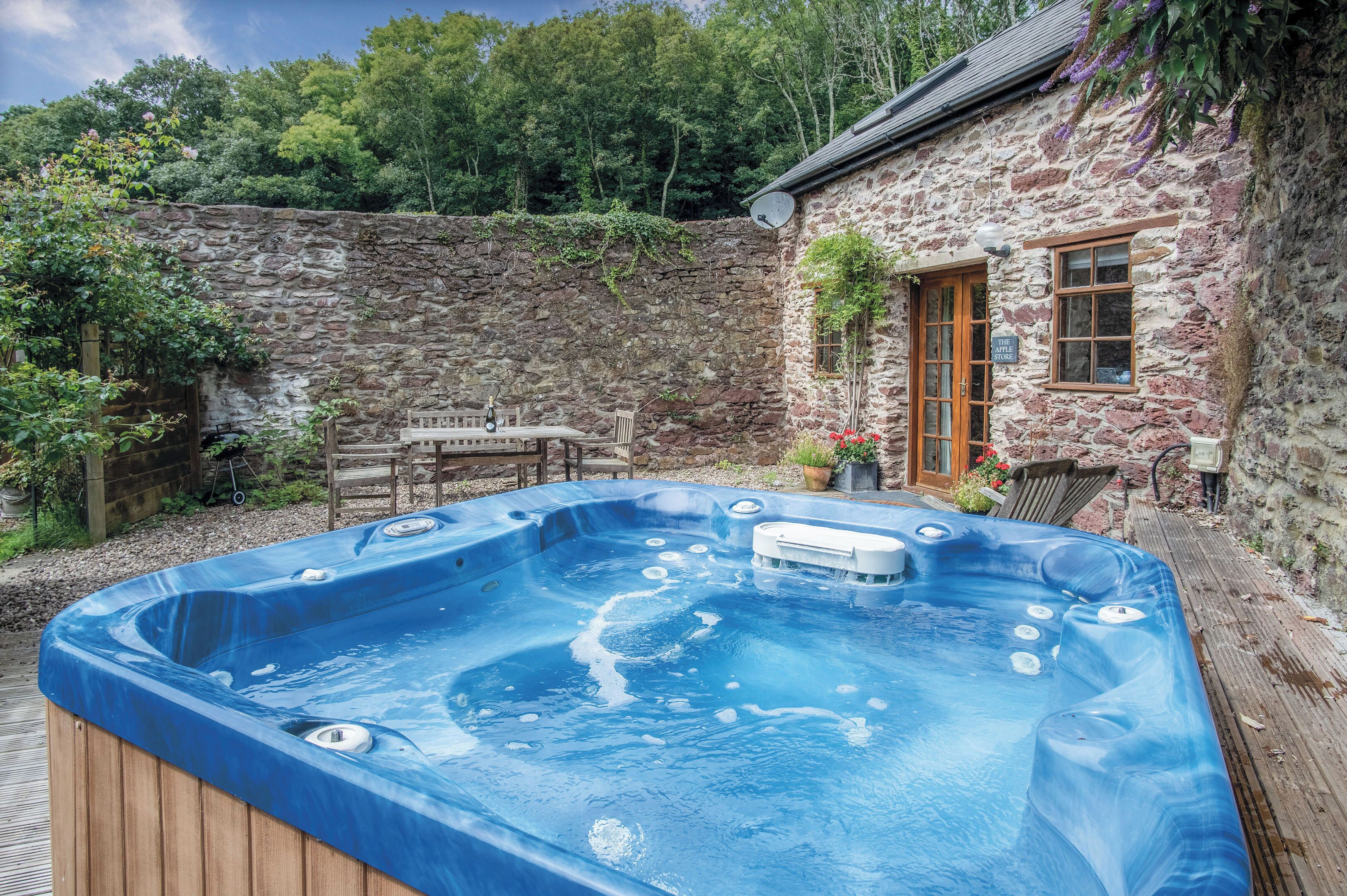 A hot tub in a stone-walled outdoor area with a wooden deck, a table, and chairs.