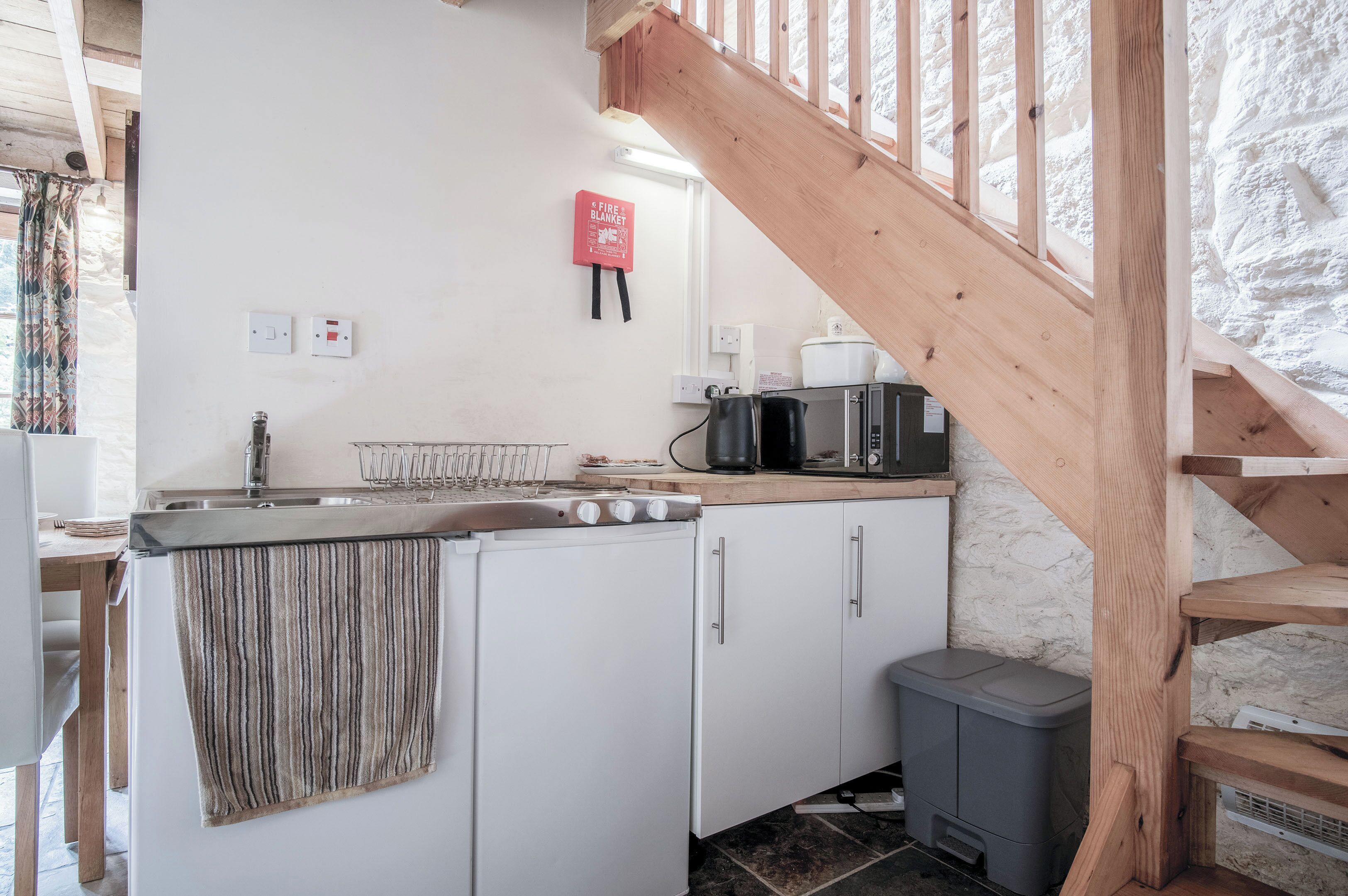 A compact kitchen with a wooden staircase, a fire blanket, and a kettle.