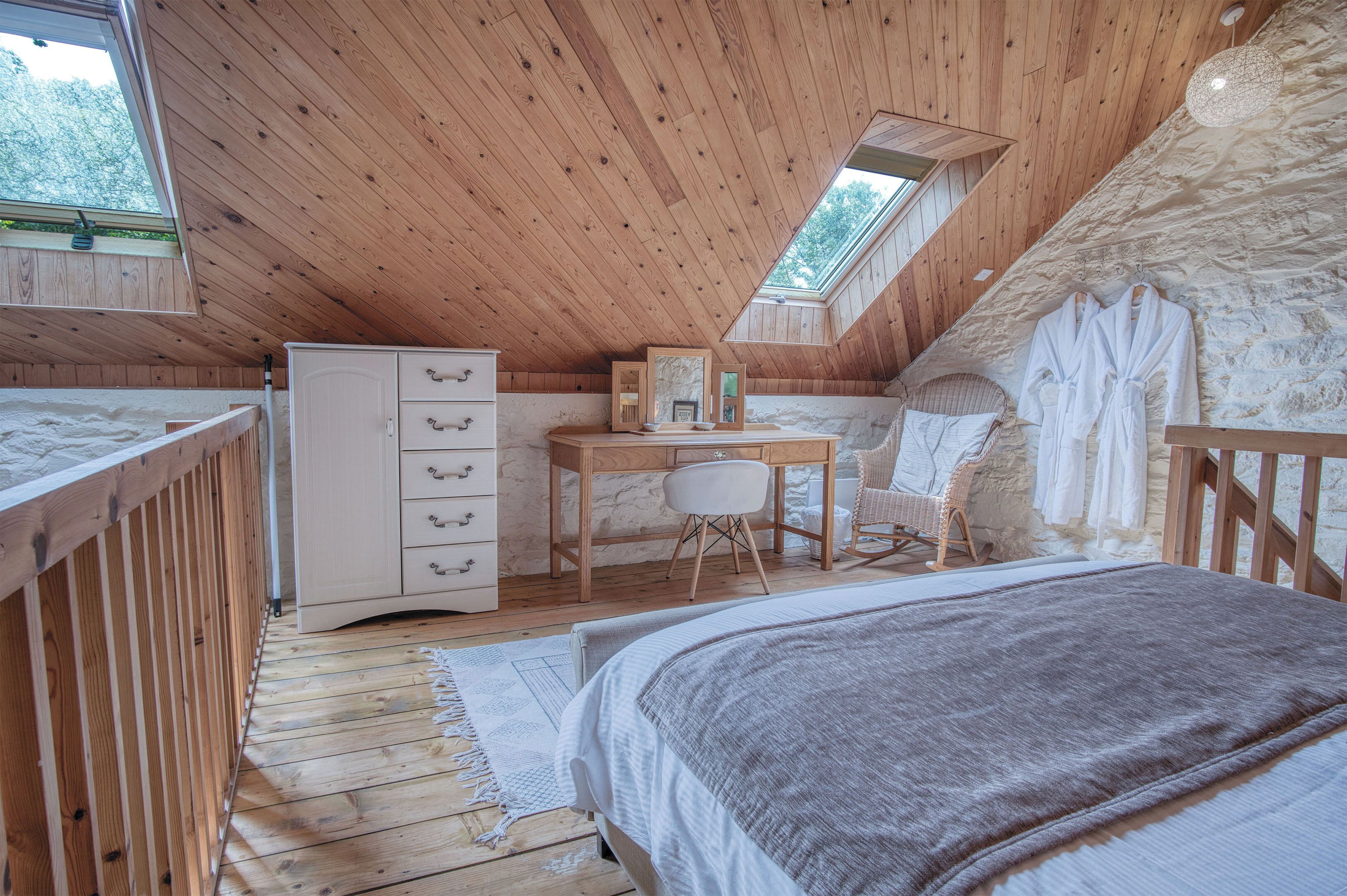 A cozy attic bedroom with a wooden ceiling, a desk with a chair, a bed with a blanket, and a wardrobe.