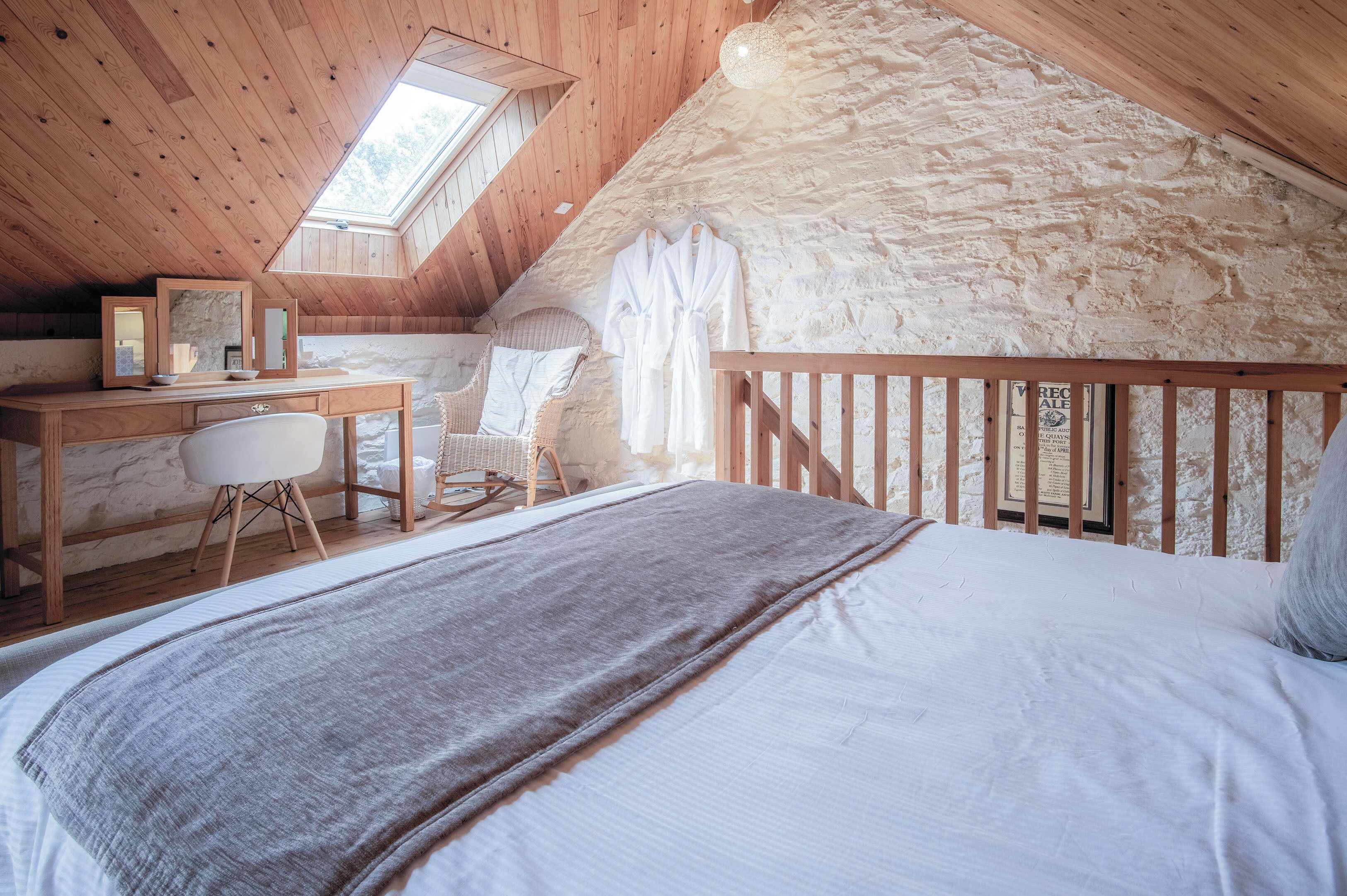 A cozy attic bedroom with a wooden desk, a comfortable chair, a bed with a gray blanket, and a stone wall.