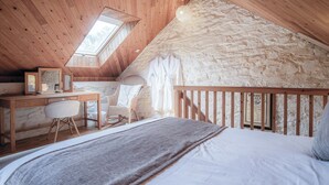 A cozy attic bedroom with a wooden desk, a comfortable chair, a bed with a gray blanket, and a stone wall.
