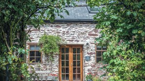 A stone building with a wooden door and windows, surrounded by greenery.