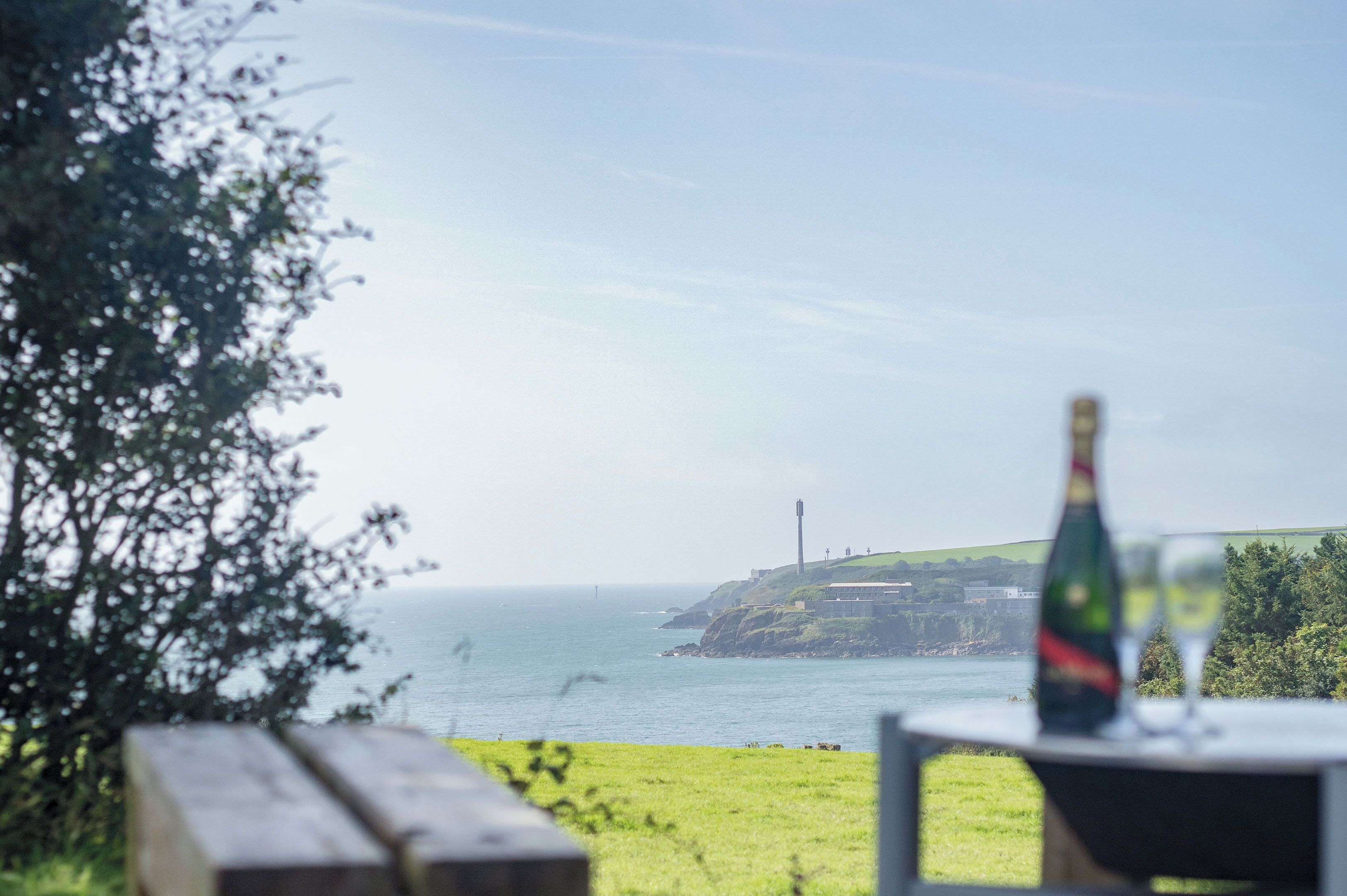 Une vue sur la côte, avec un banc en bois et une table sur laquelle se trouvent une bouteille et des verres, donnant sur la mer et une falaise lointaine parsemée de constructions.