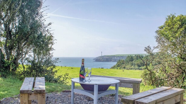 Een houten bank en tafelset buiten, met een fles en glazen op de tafel, met uitzicht op een kustlandschap.