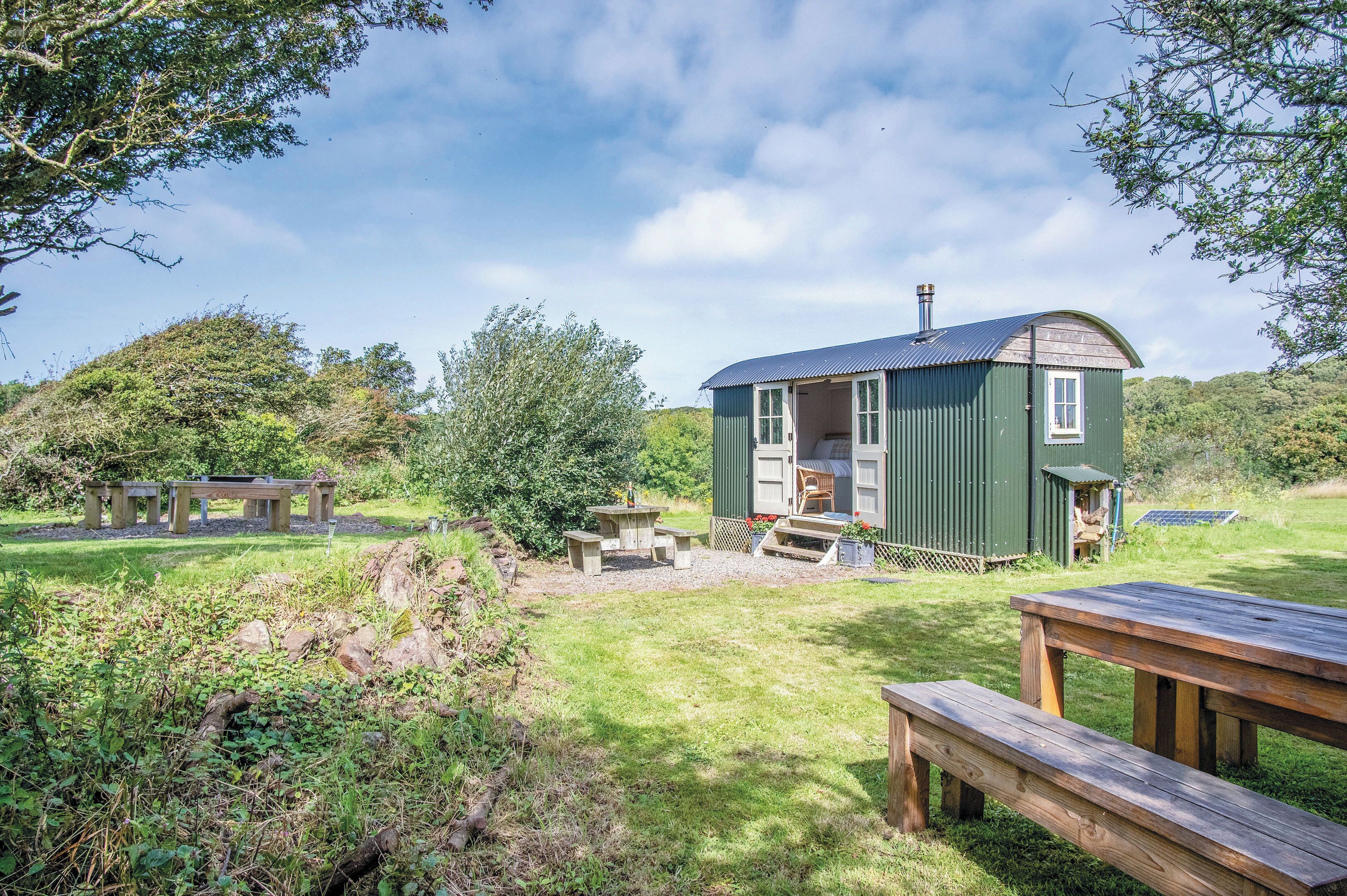 A green cottage with a door and windows, a wooden bench, and a picnic table in a grassy area with trees.