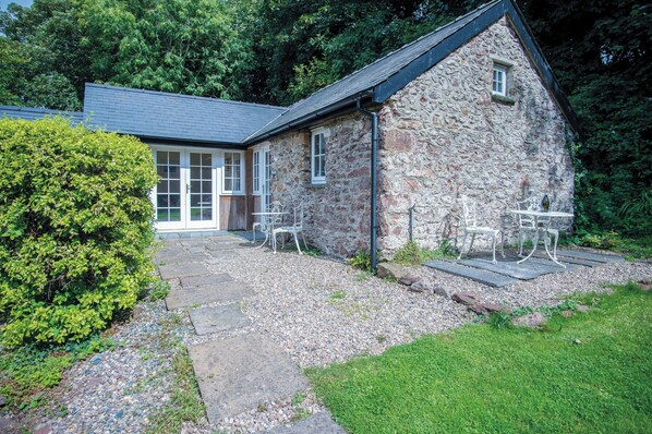 A stone building with a sloped roof, a door, and a window. There are two white chairs outside.