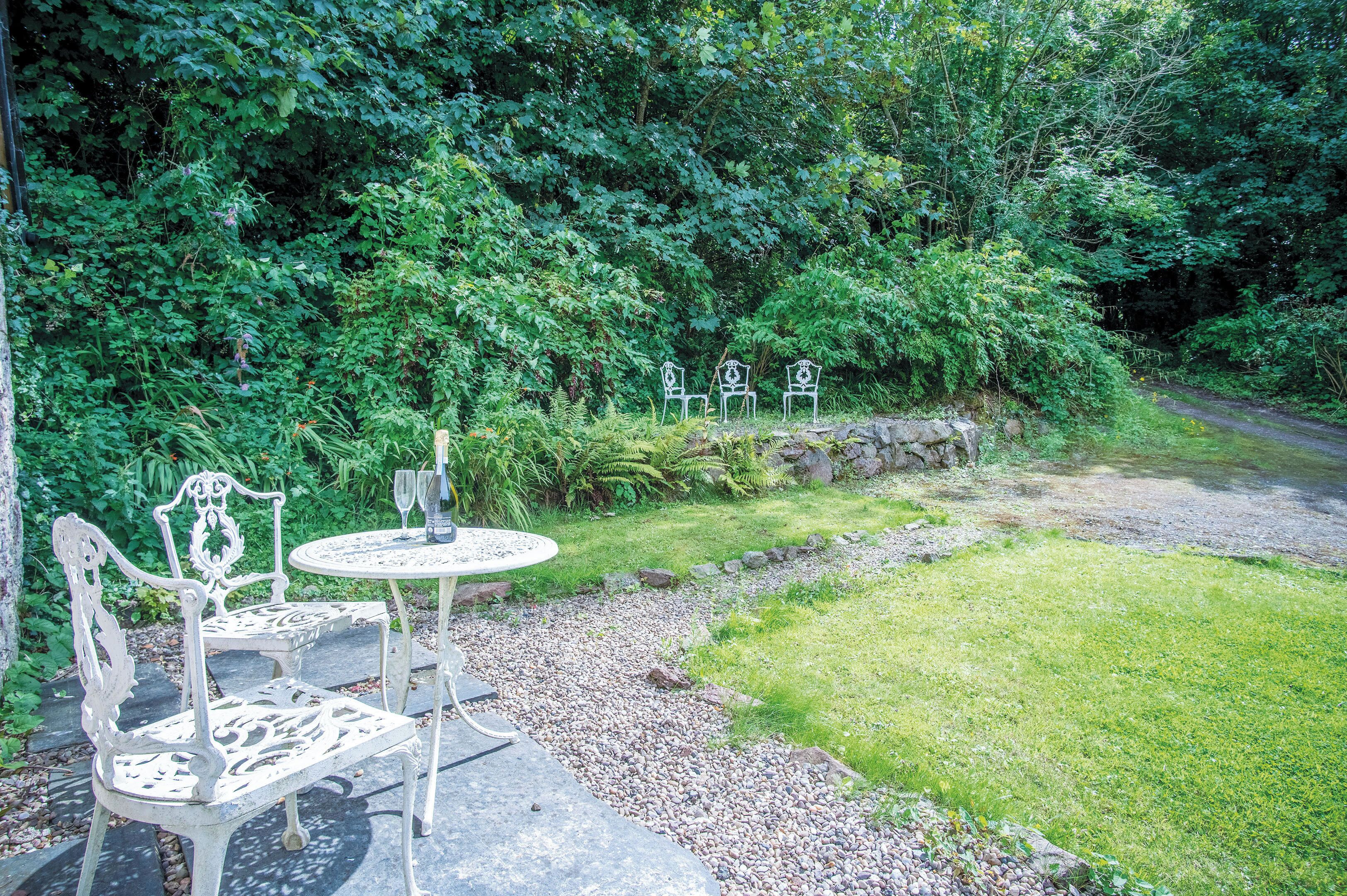 A garden with a stone table and two chairs, surrounded by lush greenery and a gravel path.