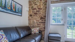Un salon avec un mur en pierre, un canapé en cuir noir, un coussin coloré, un petit pouf et un tableau représentant des voiliers.