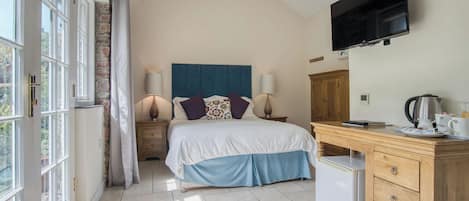 A bedroom with a bed, bedside lamps, a television, a desk with a kettle, and a refrigerator.