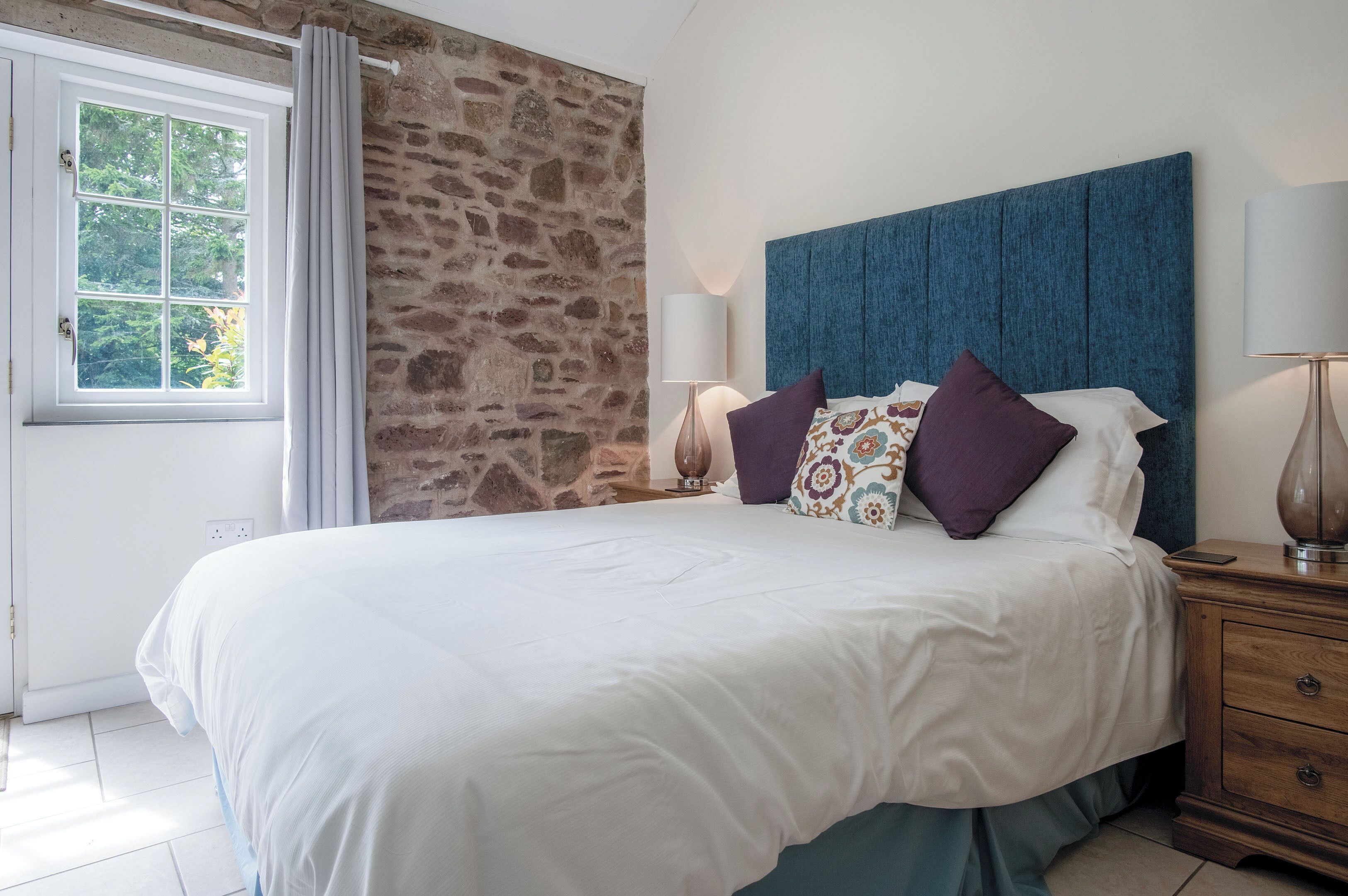 Ein Schlafzimmer mit Steinwand, einem großen Bett mit weißer Bettwäsche und vielen Kissen, einem Nachttisch aus Holz mit Lampe sowie einem Fenster mit Vorhängen.