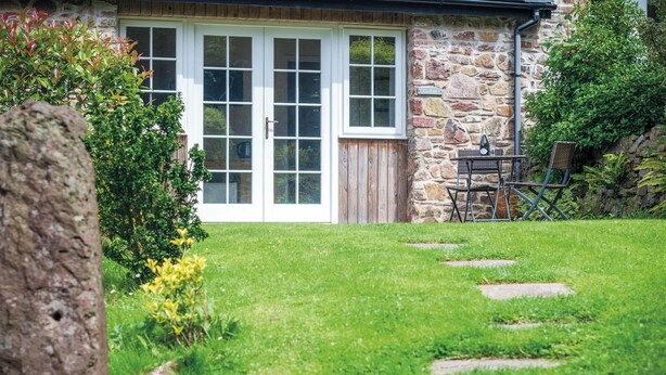 Une maison en pierre avec une porte et des fenêtres en bois, une cour verdoyante avec des dalles en pierre servant de chemin, et une table avec des chaises à l’extérieur.
