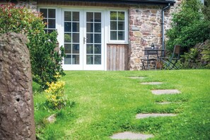 Une maison en pierre avec une porte et des fenêtres en bois, une cour verdoyante avec des dalles en pierre servant de chemin, et une table avec des chaises à l’extérieur.